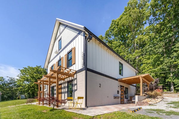Modern farmhouse with white and gray exterior, wooden pergolas, patio seating, surrounded by trees and green lawn